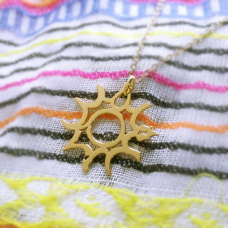 Gold sun motif necklace laying on stripe scarf. The motif is designed with Arabic word that means sun.